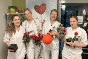 Fire personer i hvite arbeidsklær holder roser, konfektesker, blomster og en rød ballong foran en plakat med teksten ‘GiBlod.no’ og Røde Kors-logo