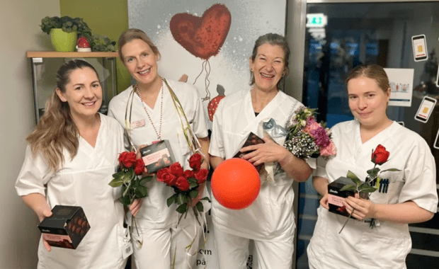 Fire personer i hvite arbeidsklær holder roser, konfektesker, blomster og en rød ballong foran en plakat med teksten ‘GiBlod.no’ og Røde Kors-logo