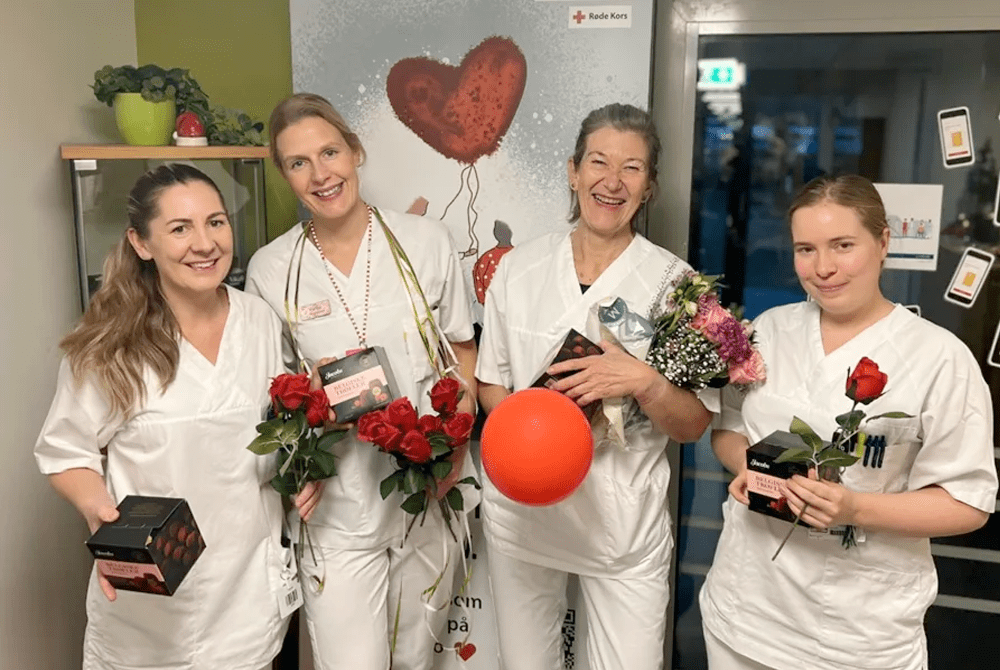 Fire personer i hvite arbeidsklær holder roser, konfektesker, blomster og en rød ballong foran en plakat med teksten ‘GiBlod.no’ og Røde Kors-logo